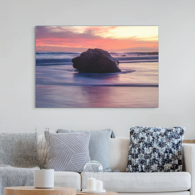 Canvas print of a beach scene with a sunset over a rock, hanging above a living room sofa.