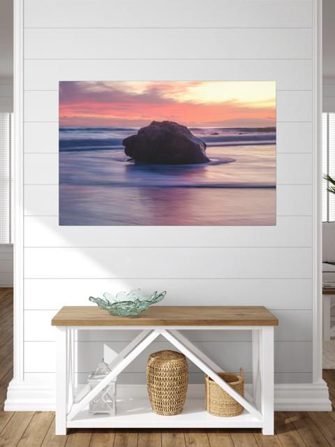 A canvas print of a large ocean rock on a wall above a wooden bench with storage baskets.