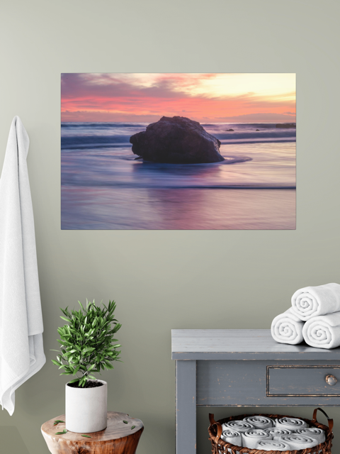 A canvas print of a large ocean rock on a wall above a table with a plant and folded towels.