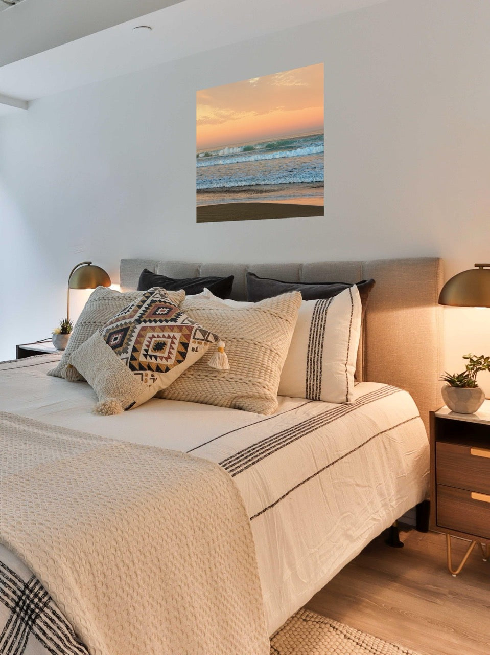 Bedroom with a bed featuring patterned pillows, a nightstand, and a canvas print of a sunrise sky over ocean waves.