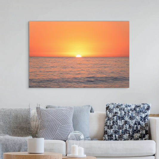 A canvas print of a sunset over the ocean on the wall over a couch.