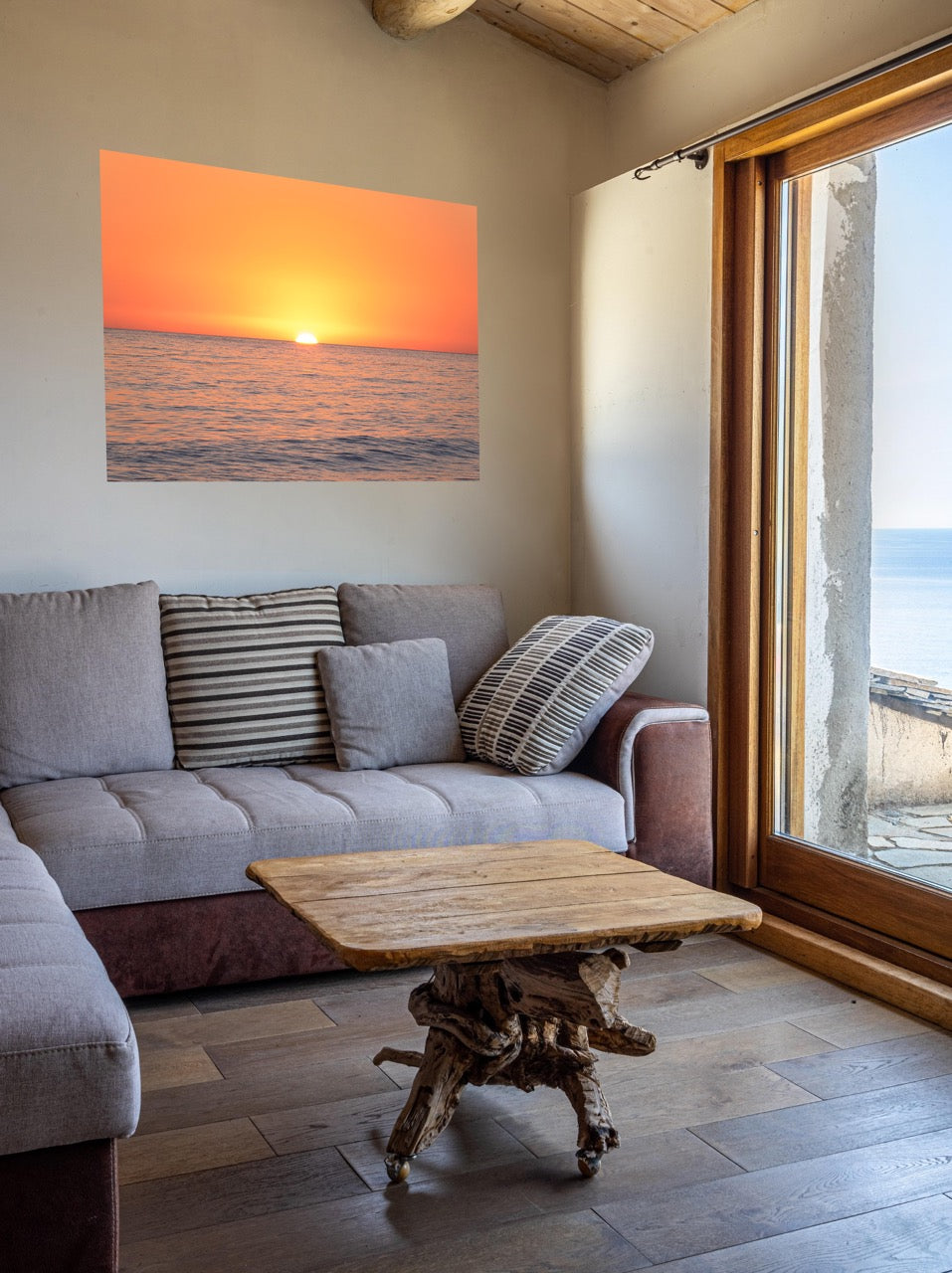 Acanvas print of a sunset over the ocean over a sectional sofa in a living room with a large window.
