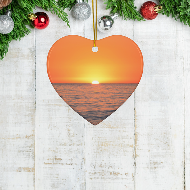 Heart-shaped ceramic Christmas ornament featuring a minimalist photograph of a vibrant orange and amber sunset sky over a calm Pacific Ocean on a wooden surface with Christmas decorations.