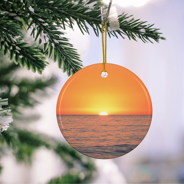 Ceramic Christmas ornament featuring a minimalist photograph of a vibrant orange and amber sunset sky over a calm Pacific Ocean on a tree branch