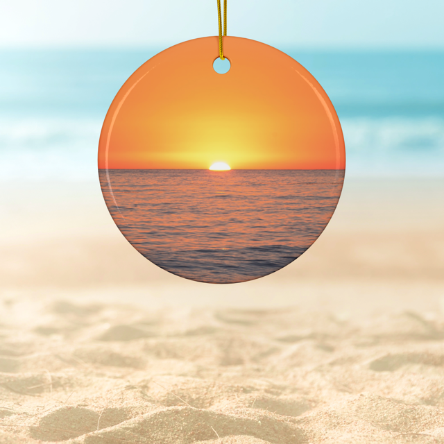 Ceramic Christmas ornament featuring a minimalist photograph of a vibrant orange and amber sunset sky over a calm Pacific Ocean on a sandy background