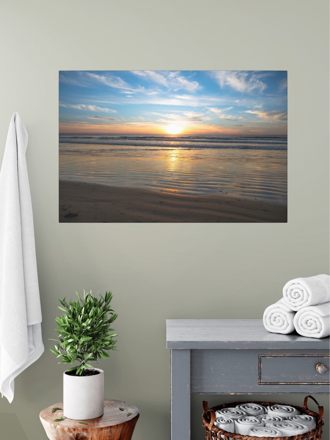 California beach sunset over the ocean with golden sand ripples canvas print on a wall above a table with a plant and folded towels.