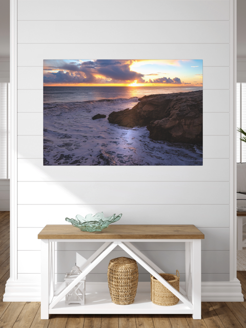 California coastal sunset after rain storm canvas print on a wall, with a wooden bench and decorative items below.