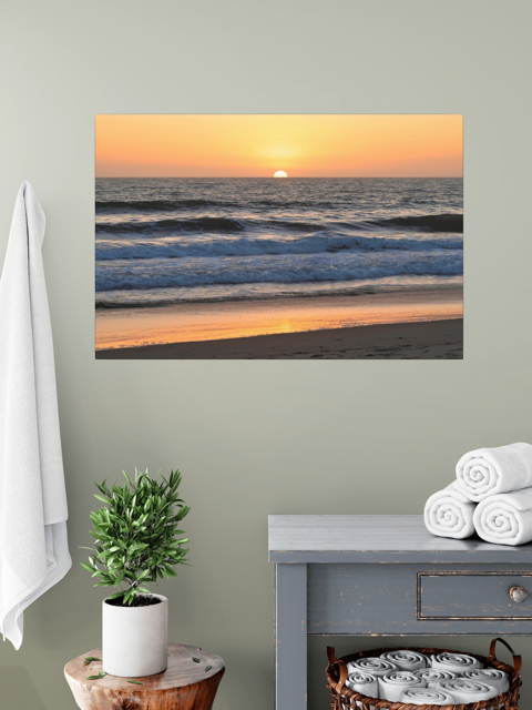 Pacific Ocean sunset and waves on beach canvas print on a wall above a plant and shelves.