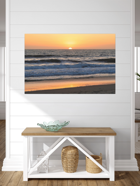 Pacific Ocean sunset and waves on beach canvas print on a wall above a wooden bench with storage.
