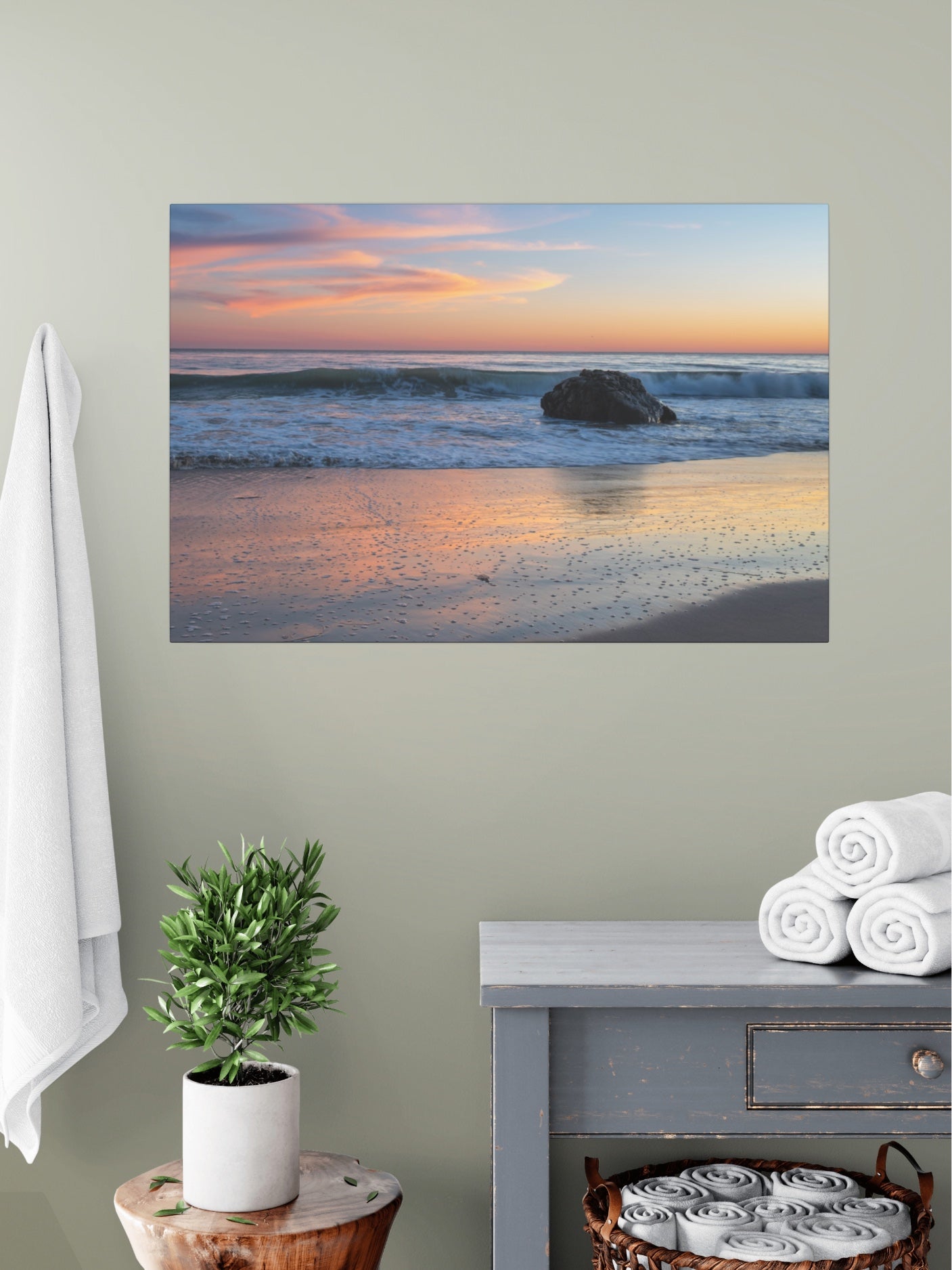 Pastel beach twilight canvas print with pink clouds reflecting on wet sand on a wall above a table with a plant and folded towels.
