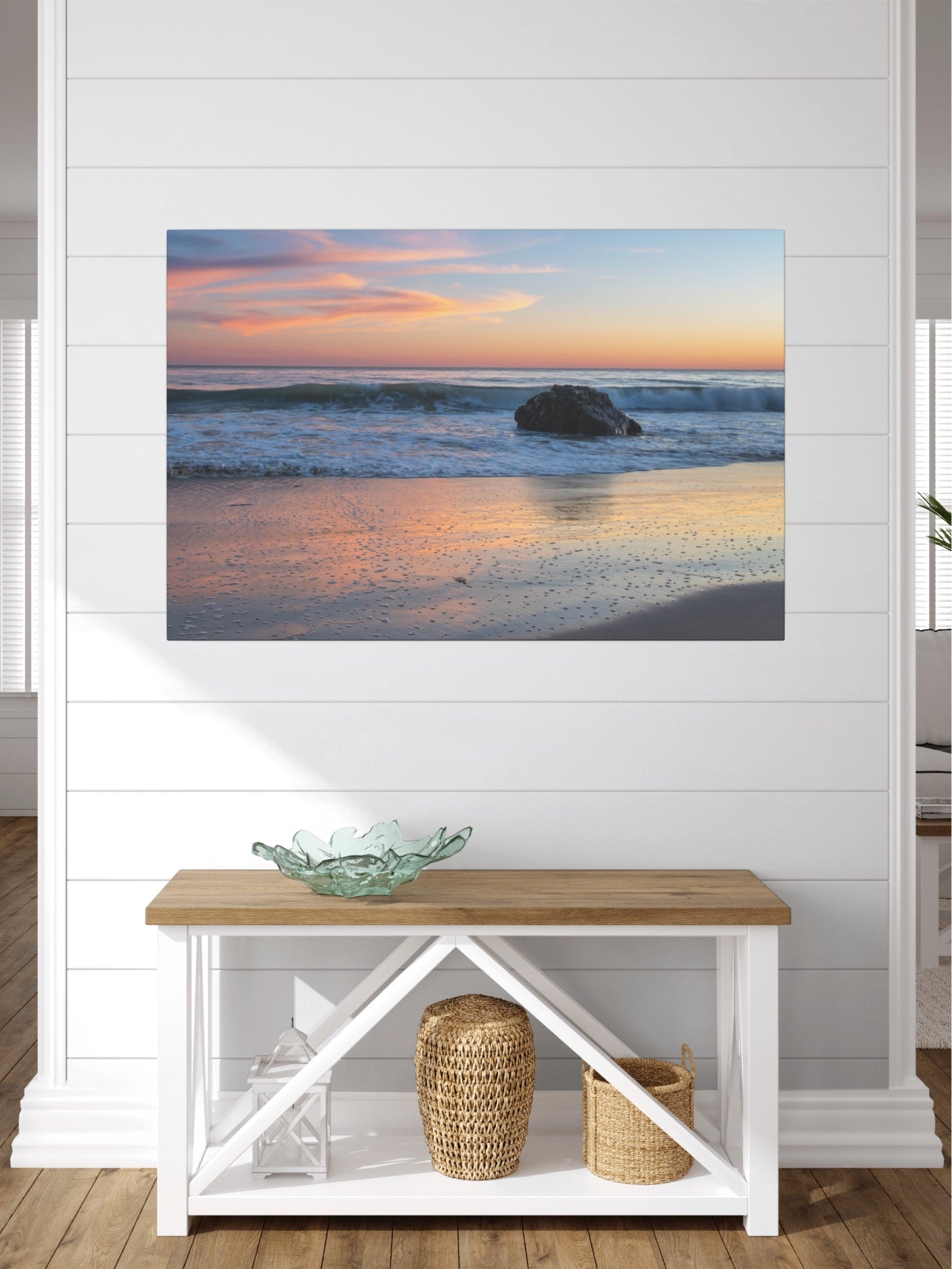Pastel beach twilight canvas print with pink clouds reflecting on wet sand on a wall above a wooden bench with a glass bowl and baskets.