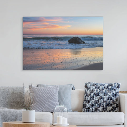 Pastel beach twilight canvas print with pink clouds reflecting on wet sand on a wall above a sofa with decorative pillows.