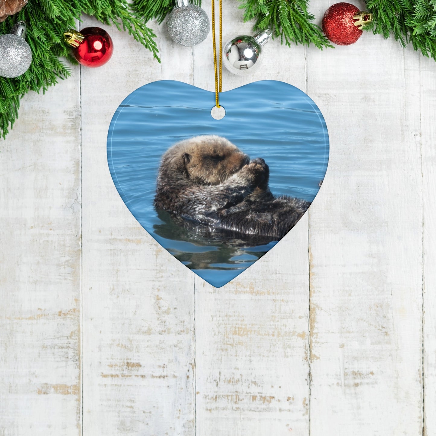 Heart-shaped ceramic Christmas ornament featuring a peaceful photograph of a sea otter with its eyes closed, on a wooden surface with Christmas decorations.