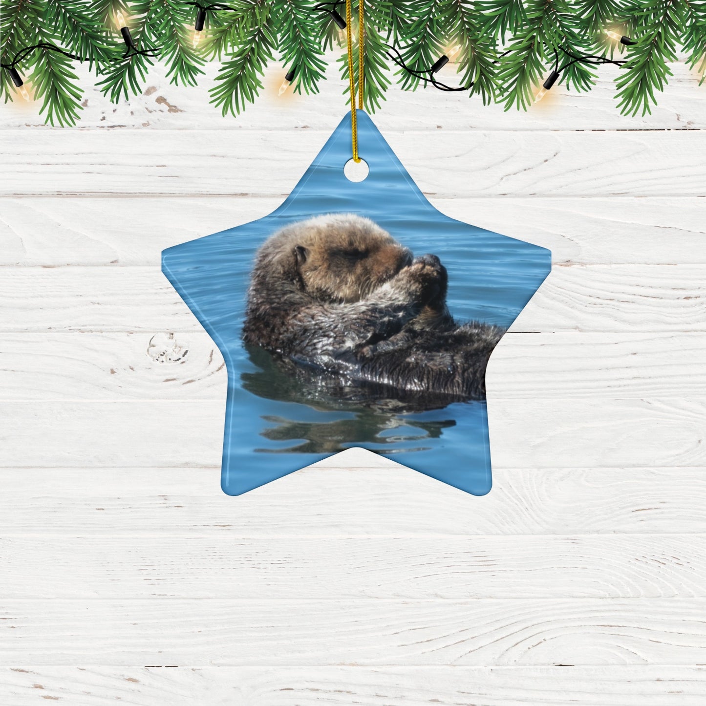 Star-shaped ceramic Christmas ornament featuring a peaceful photograph of a sea otter with its eyes closed, on a white wooden background.