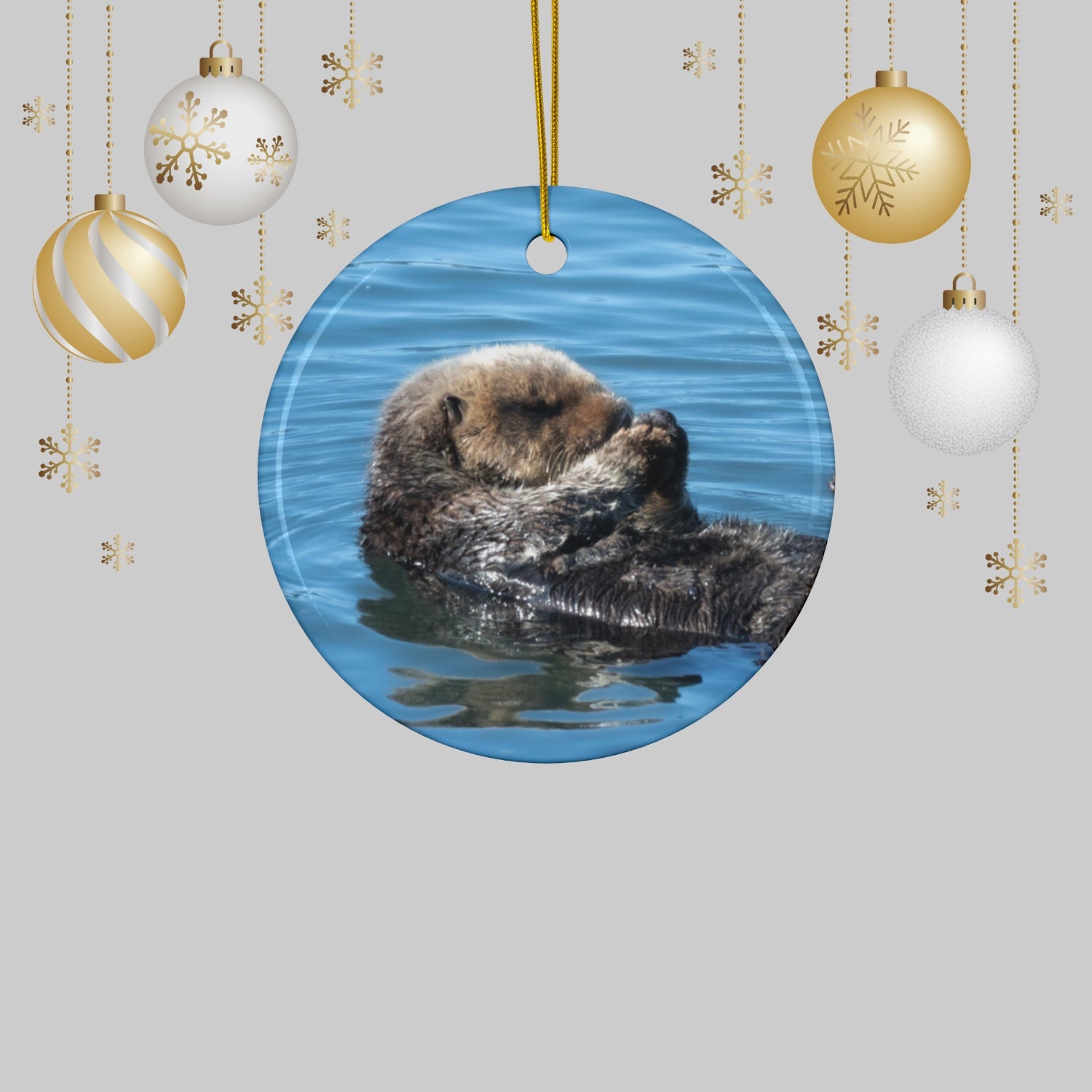 Ceramic Christmas ornament featuring a peaceful photograph of a sea otter with its eyes closed, surrounded by Christmas ornaments on a light gray background