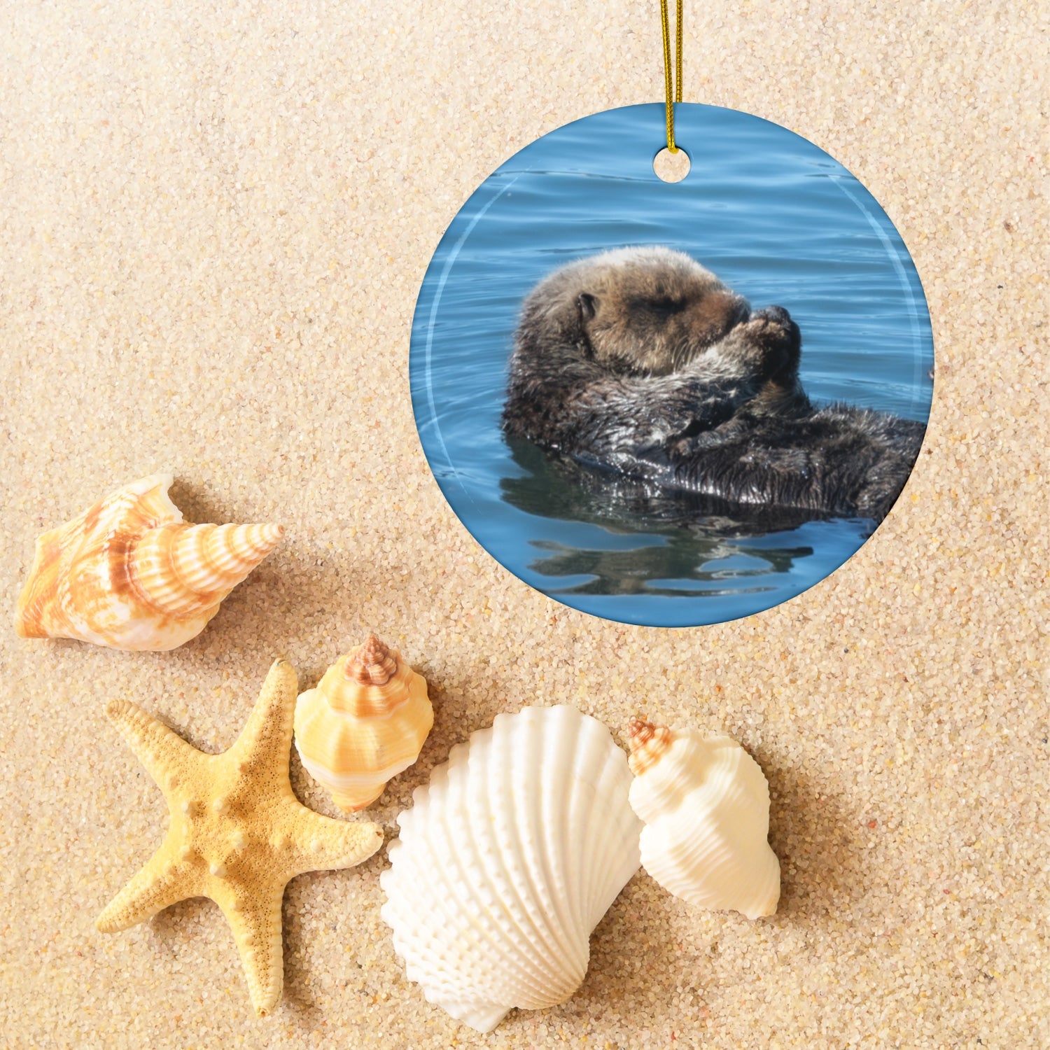 Ceramic Christmas ornament featuring a peaceful photograph of a sea otter with its eyes closed, on a sandy background with shells and a starfish.