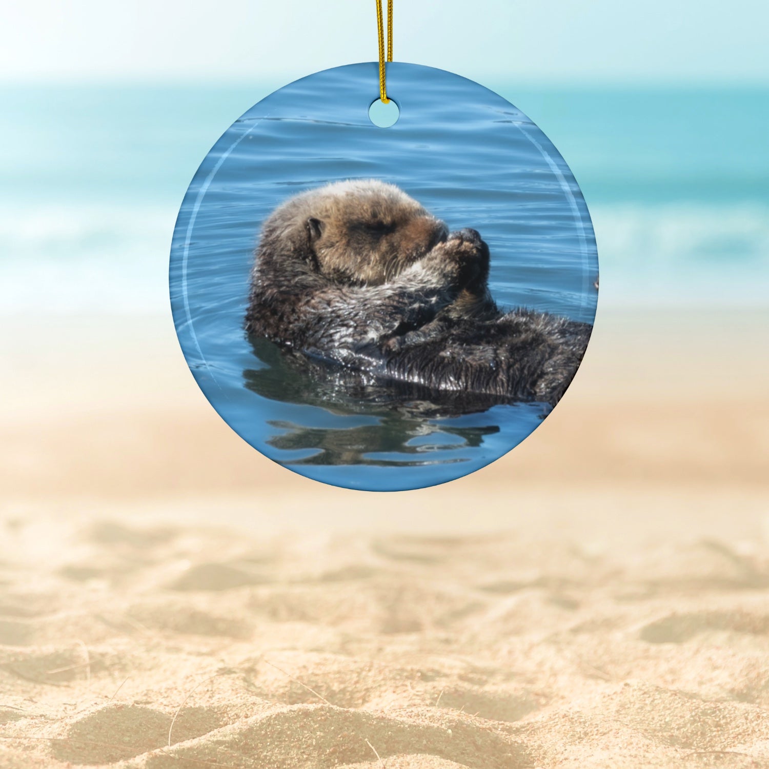 Ceramic Christmas ornament featuring a peaceful photograph of a sea otter with its eyes closed, hanging against a beach background.