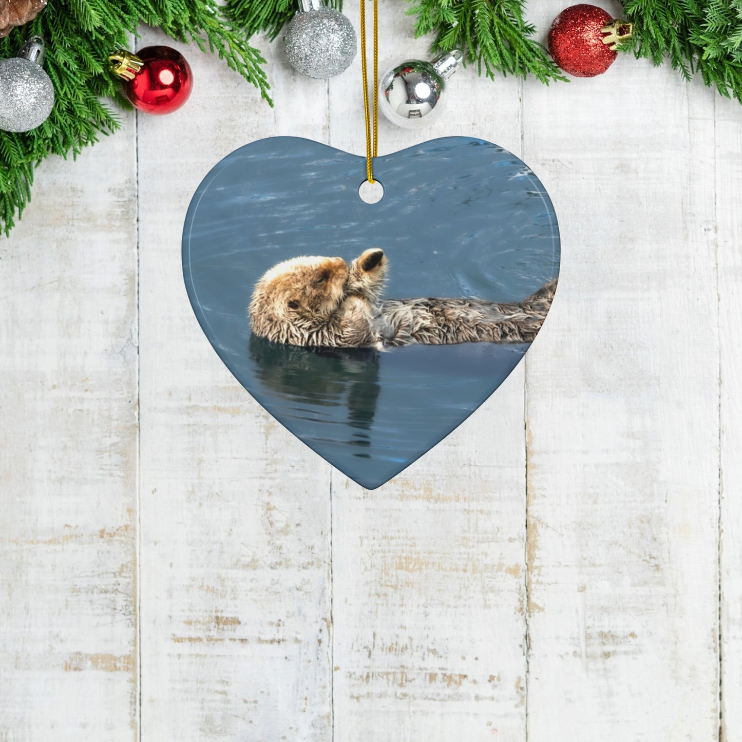 Heart-shaped ornament featuring a professional photograph of a sea otter floating on its back in the blue waters of the Pacific Ocean on a wooden surface with Christmas decorations.