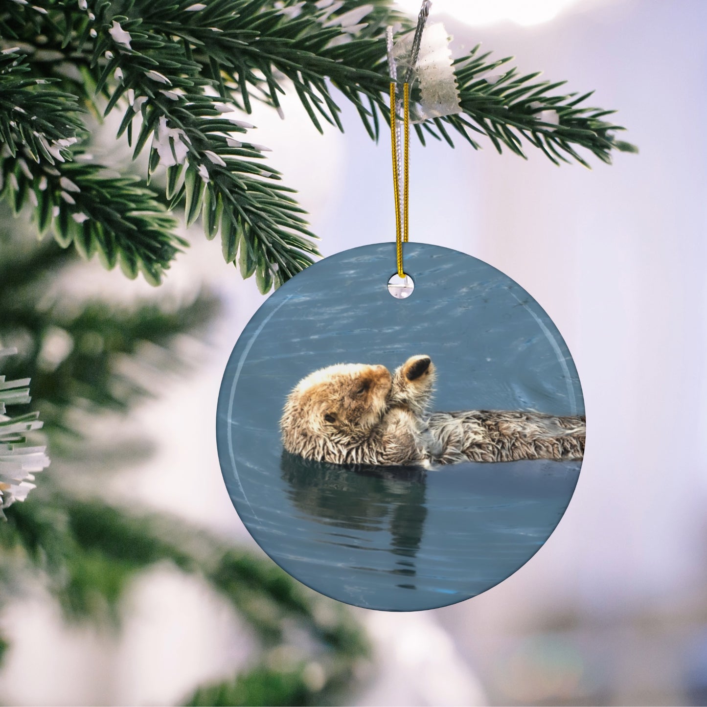 Round ornament featuring a professional photograph of a sea otter floating on its back in the blue waters of the Pacific Ocean hanging on a tree branch.