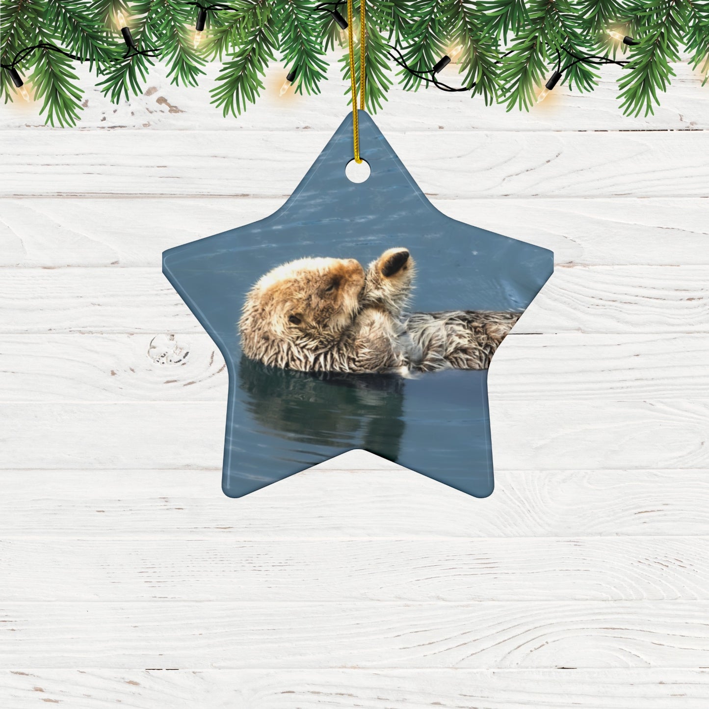 Star-shaped Christmas ornament  featuring a professional photograph of a sea otter floating on its back in the blue waters of the Pacific Ocean hanging on a wooden surface with greenery.