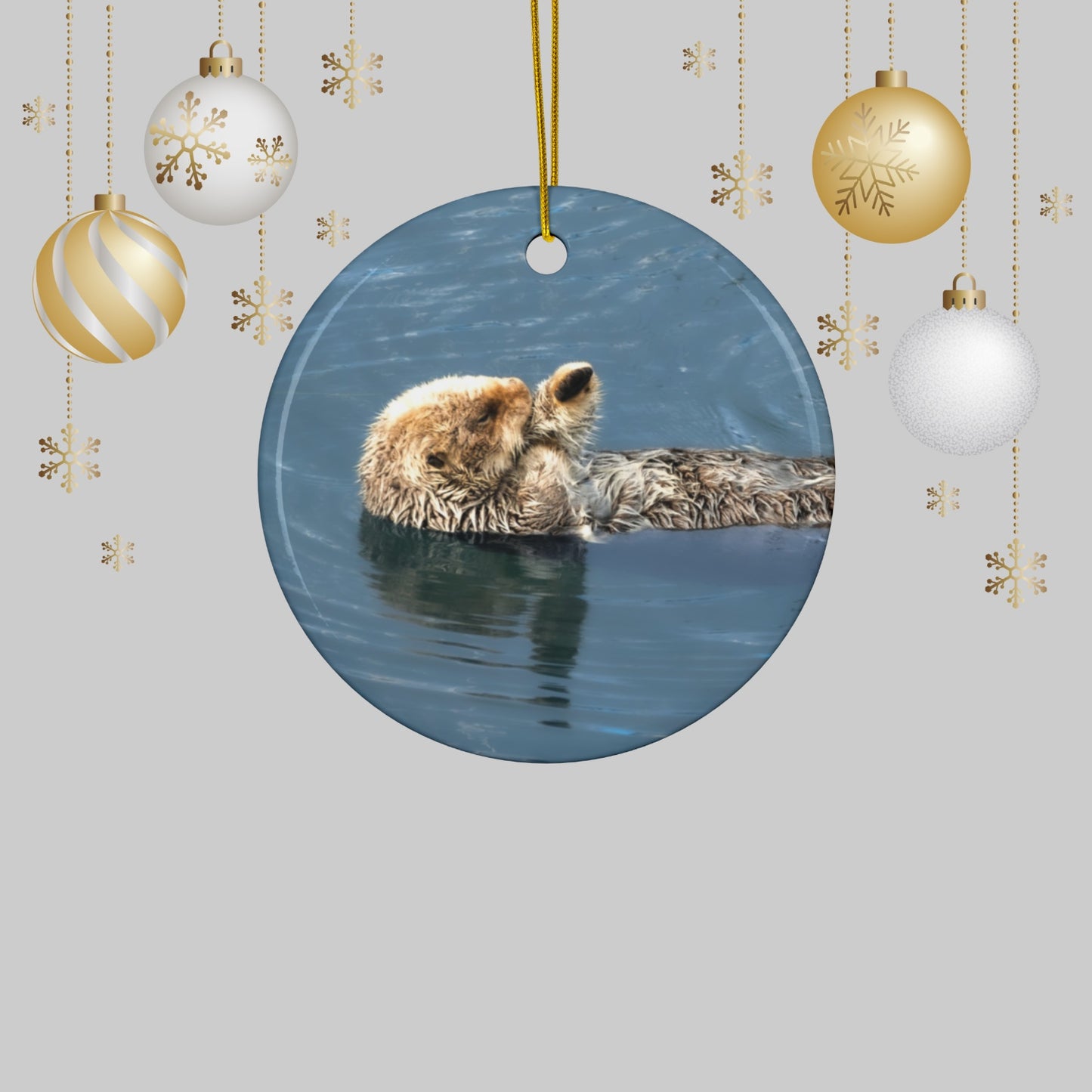 Round ornament featuring a professional photograph of a sea otter floating on its back in the blue waters of the Pacific Ocean, surrounded by Christmas ornaments and snowflakes.