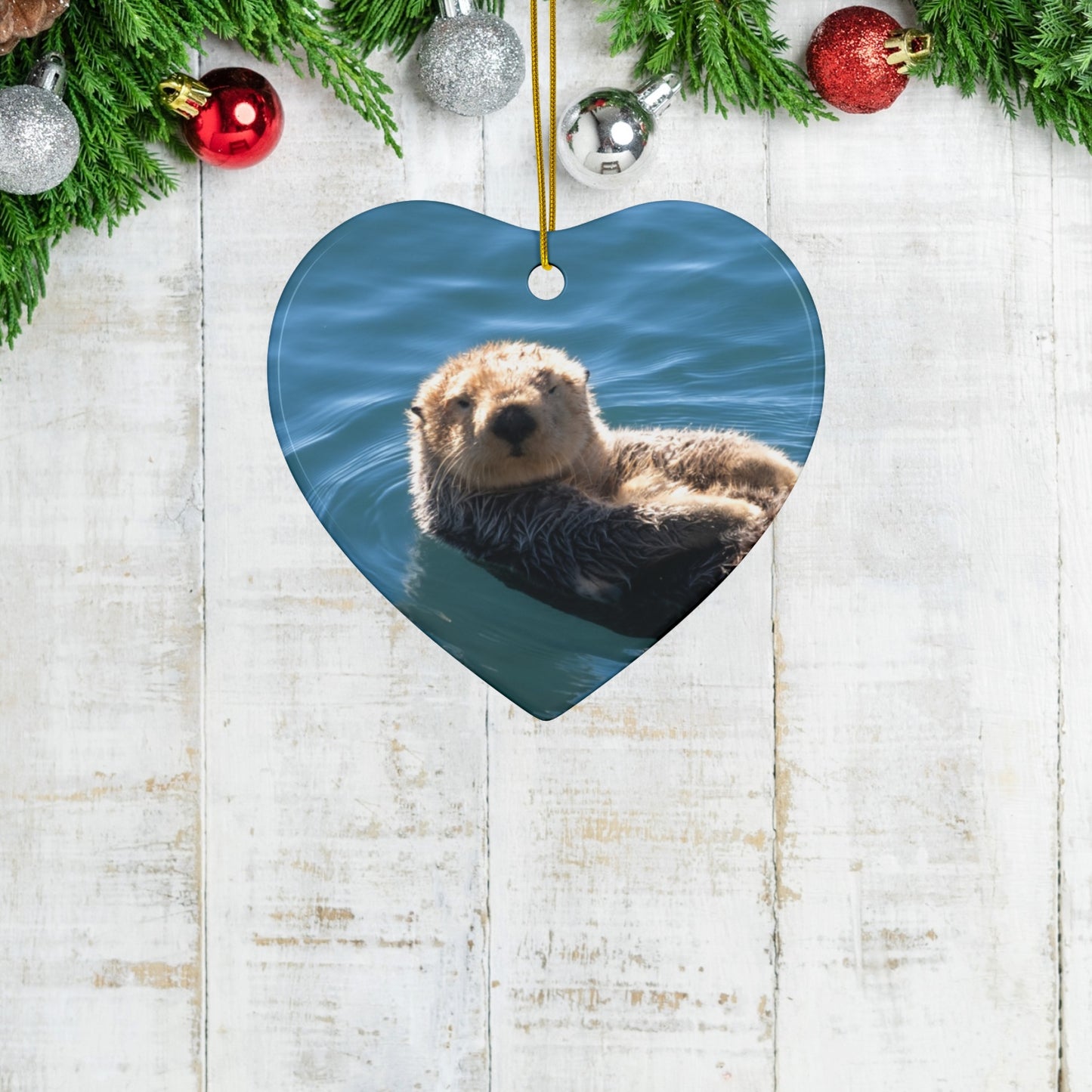 Heart-shaped ceramic Christmas ornament featuring a close-up photograph of a sea otter floating in the calm, blue waters of Morro Bay with golden light on its fur, on a wooden surface with Christmas decorations.