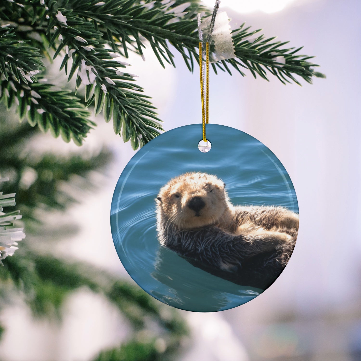 Ceramic Christmas ornament featuring a close-up photograph of a sea otter floating in the calm, blue waters of Morro Bay with golden light on its fur, hanging on a Christmas tree branch.