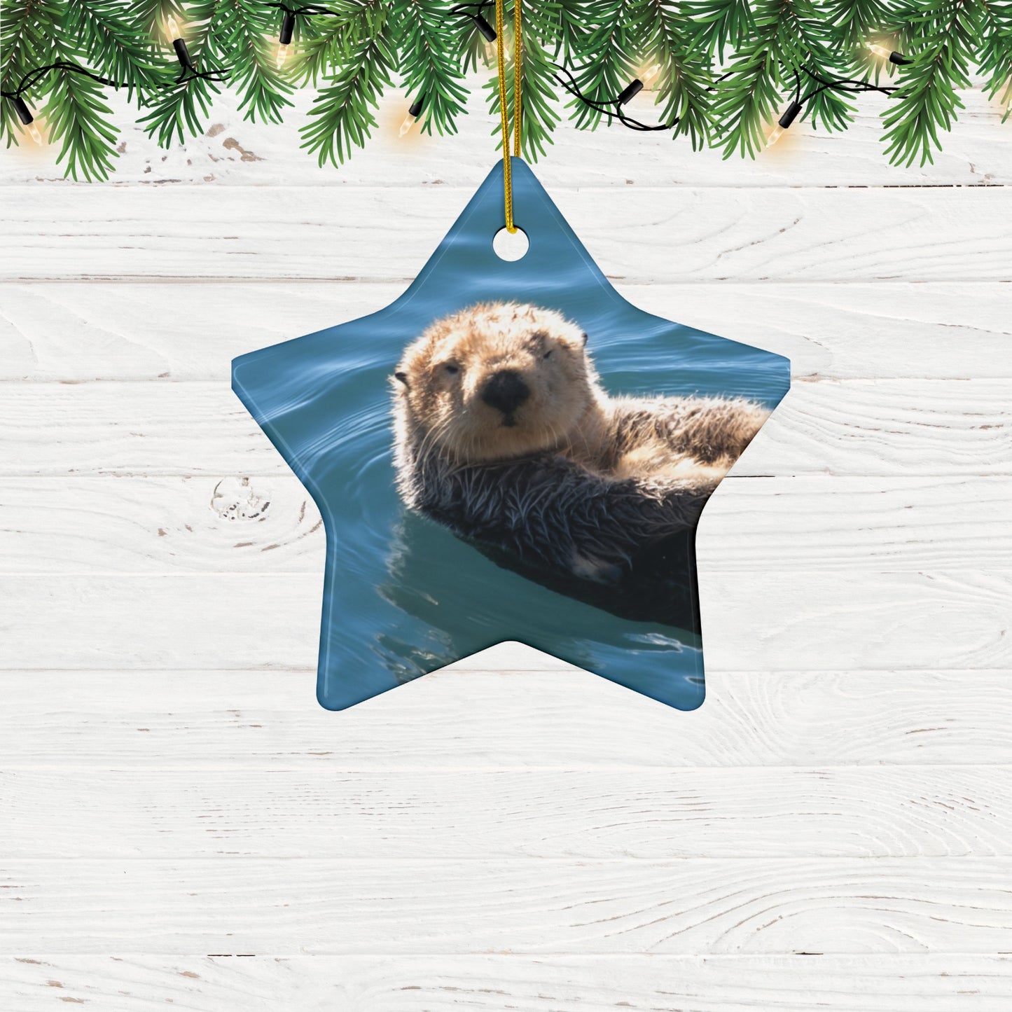 Star-shaped ceramic Christmas ornament featuring a close-up photograph of a sea otter floating in the calm, blue waters of Morro Bay with golden light on its fur, hanging from Christmas tree