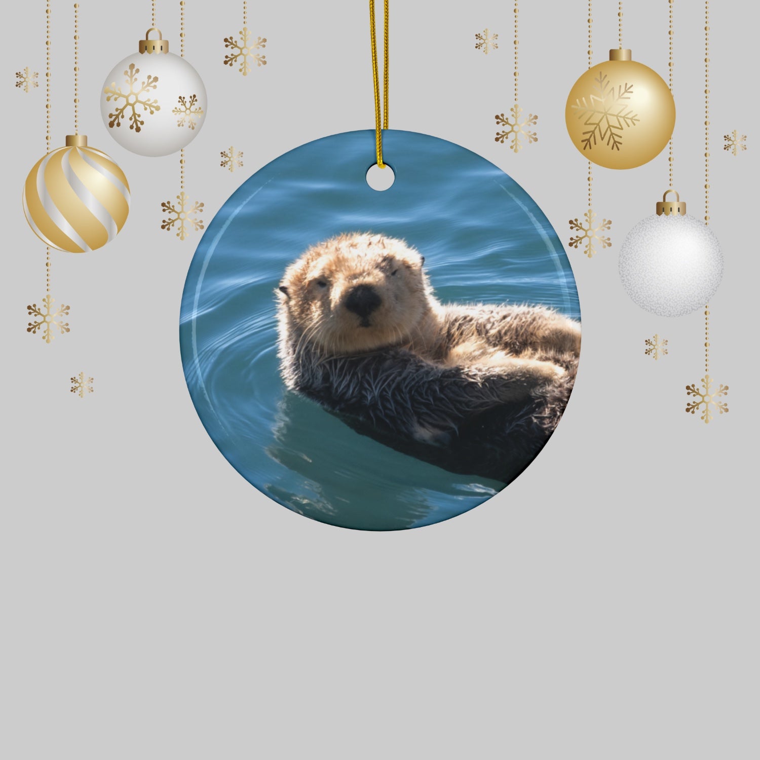 Ceramic Christmas ornament featuring a close-up photograph of a sea otter floating in the calm, blue waters of Morro Bay with golden light on its fur, surrounded by Christmas ornaments on a light gray background
