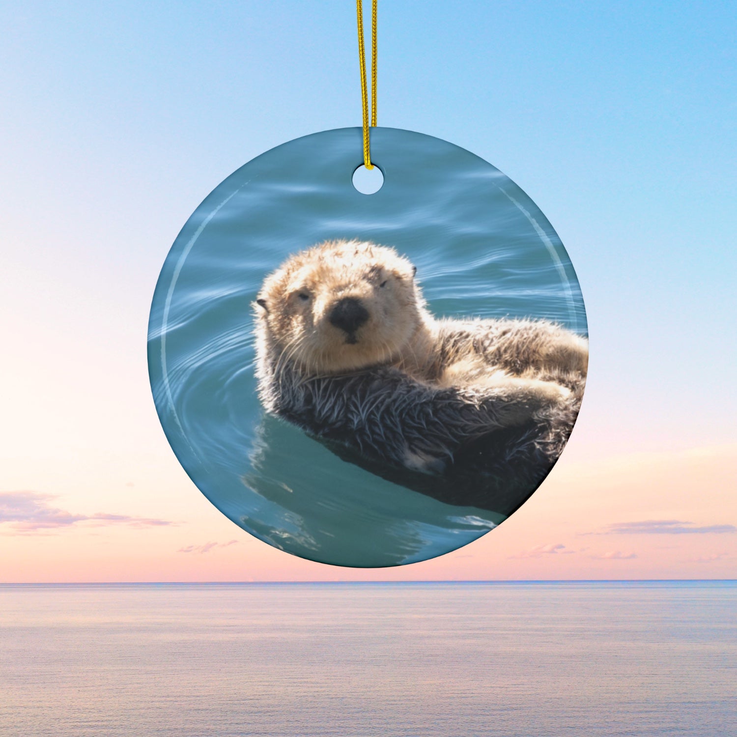 Ceramic Christmas ornament featuring a close-up photograph of a sea otter floating in the calm, blue waters of Morro Bay with golden light on its fur, against a blue sky and pink sunset background