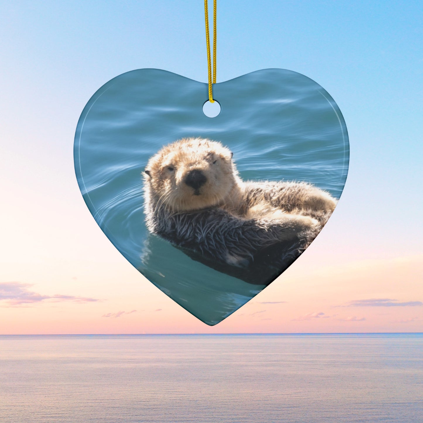 Heart-shaped ceramic Christmas ornament featuring a close-up photograph of a sea otter floating in the calm, blue waters of Morro Bay with golden light on its fur.