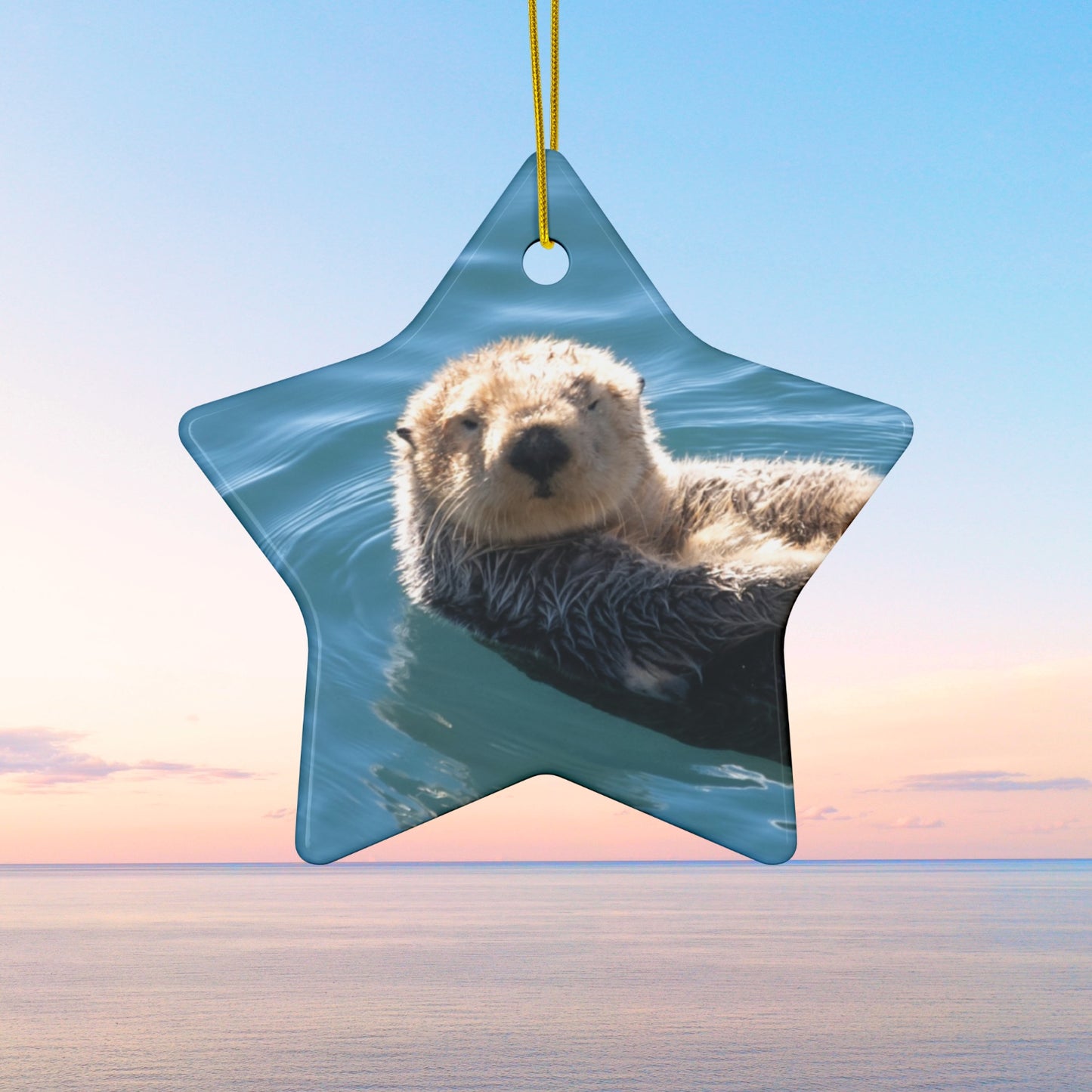 Star-shaped ceramic Christmas ornament featuring a close-up photograph of a sea otter floating in the calm, blue waters of Morro Bay with golden light on its fur.