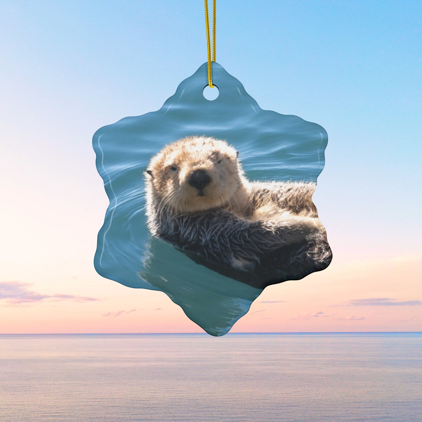 Star-shaped ceramic Christmas ornament featuring a close-up photograph of a sea otter floating in the calm, blue waters of Morro Bay with golden light on its fur, against a beach sunset background