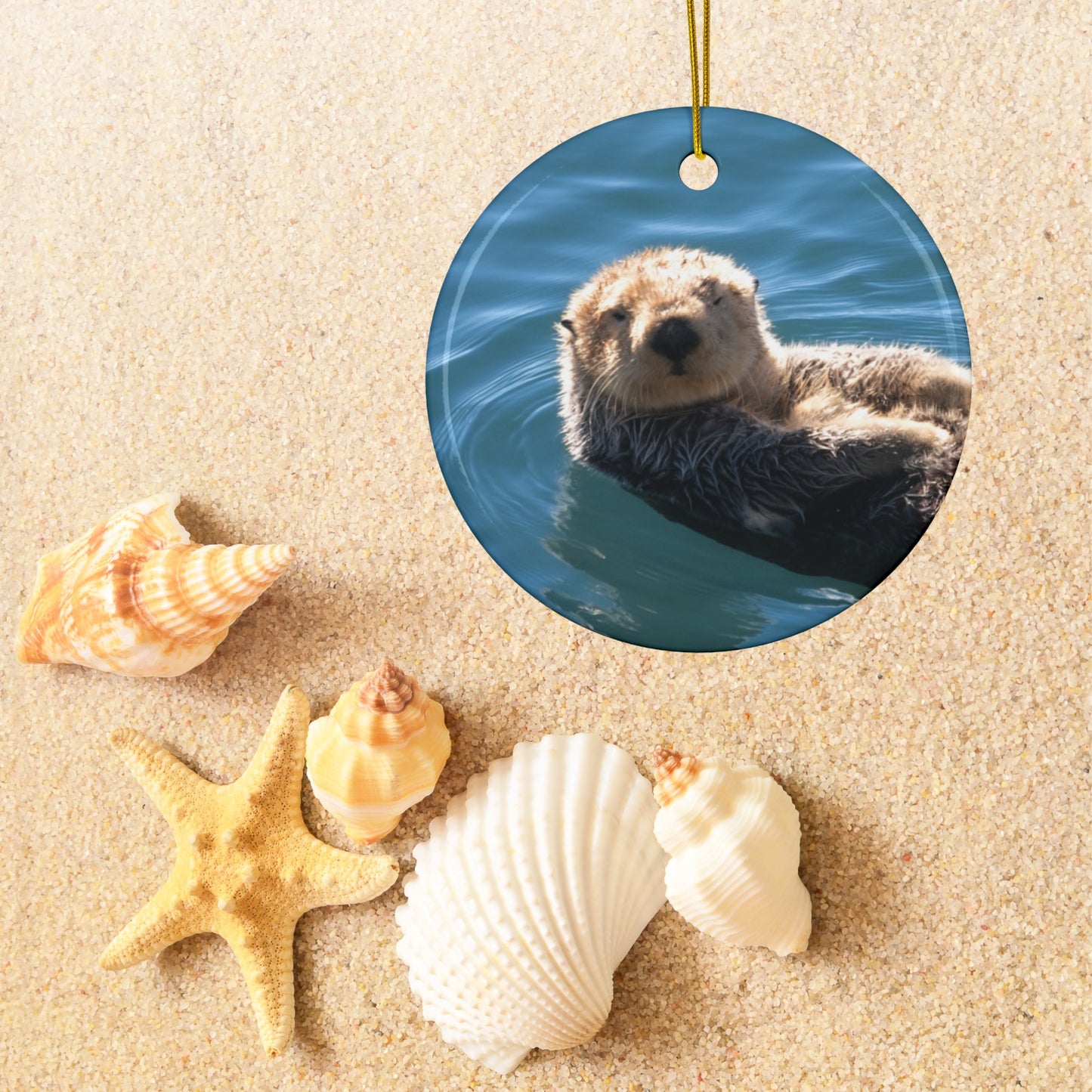 Ceramic Christmas ornament featuring a close-up photograph of a sea otter floating in the calm, blue waters of Morro Bay with golden light on its fur,  surrounded by seashells on sand