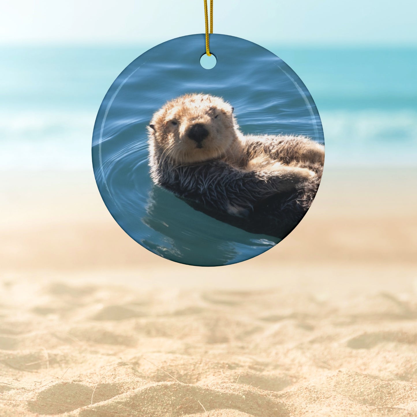 Ceramic Christmas ornament featuring a close-up photograph of a sea otter floating in the calm, blue waters of Morro Bay with golden light on its fur, hanging against a beach background.