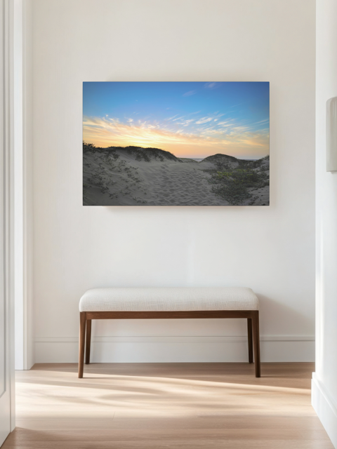 Canvas print of a beach sunset over the sand dunes on a wall above a wooden bench in a room with light wood flooring.