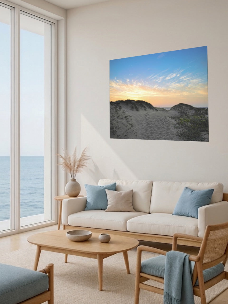Living room with a large window view of the ocean, a beige sofa with blue cushions, and a canvas print of a beach sunset over the sand dunes.