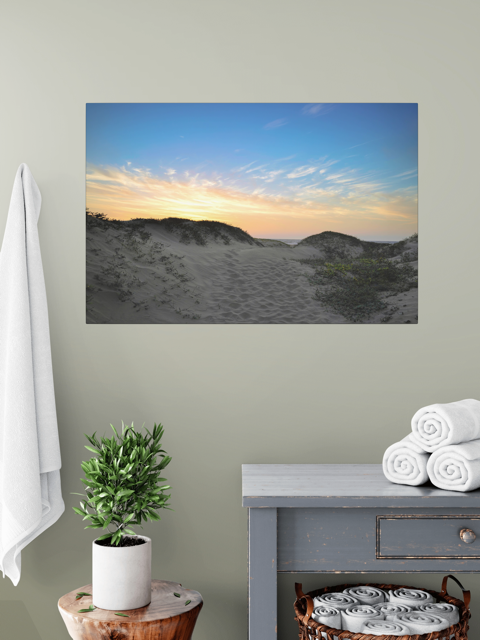 Canvas print of a beach sunset over the sand dunes over a gray table, next to a plant and folded towels on a shelf.