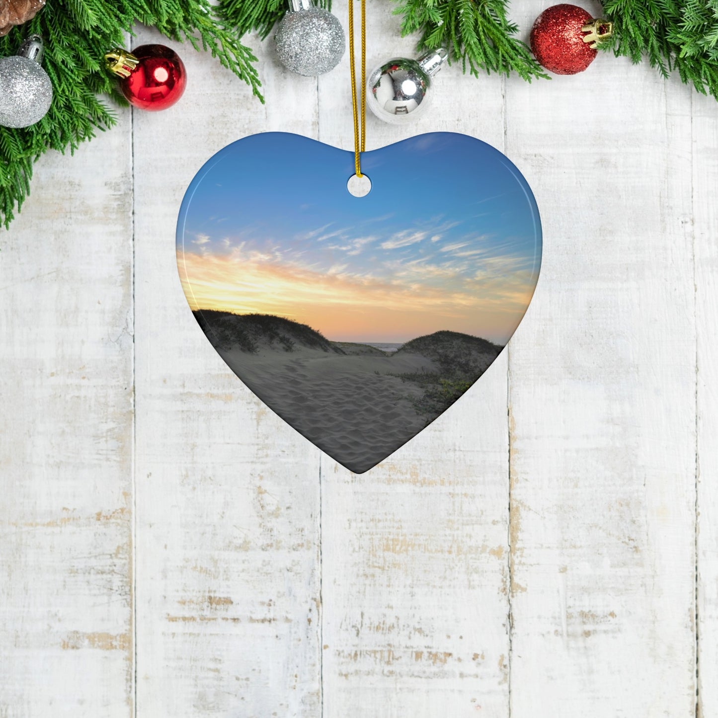 Heart-shaped ceramic holiday ornament featuring a landscape photograph of a California beach at sunset with rolling sand dunes, footprints, and gold-streaked clouds. on a wooden surface with Christmas decorations.