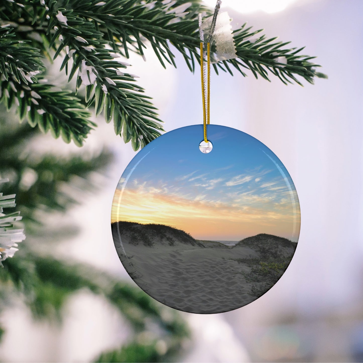 Ceramic holiday ornament featuring a landscape photograph of a California beach at sunset with rolling sand dunes, footprints, and gold-streaked clouds hanging on a Christmas tree branch.
