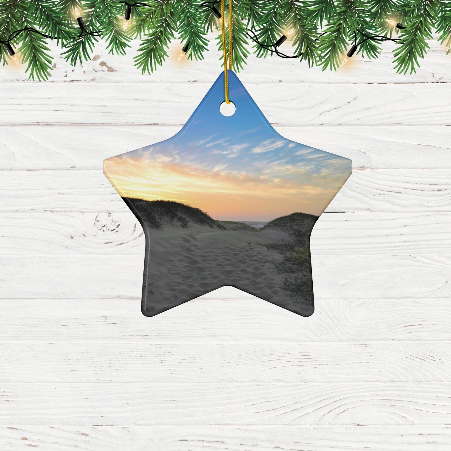Star-shaped ceramic holiday ornament featuring a landscape photograph of a California beach at sunset with rolling sand dunes, footprints, and gold-streaked clouds hanging on a wooden surface with greenery.