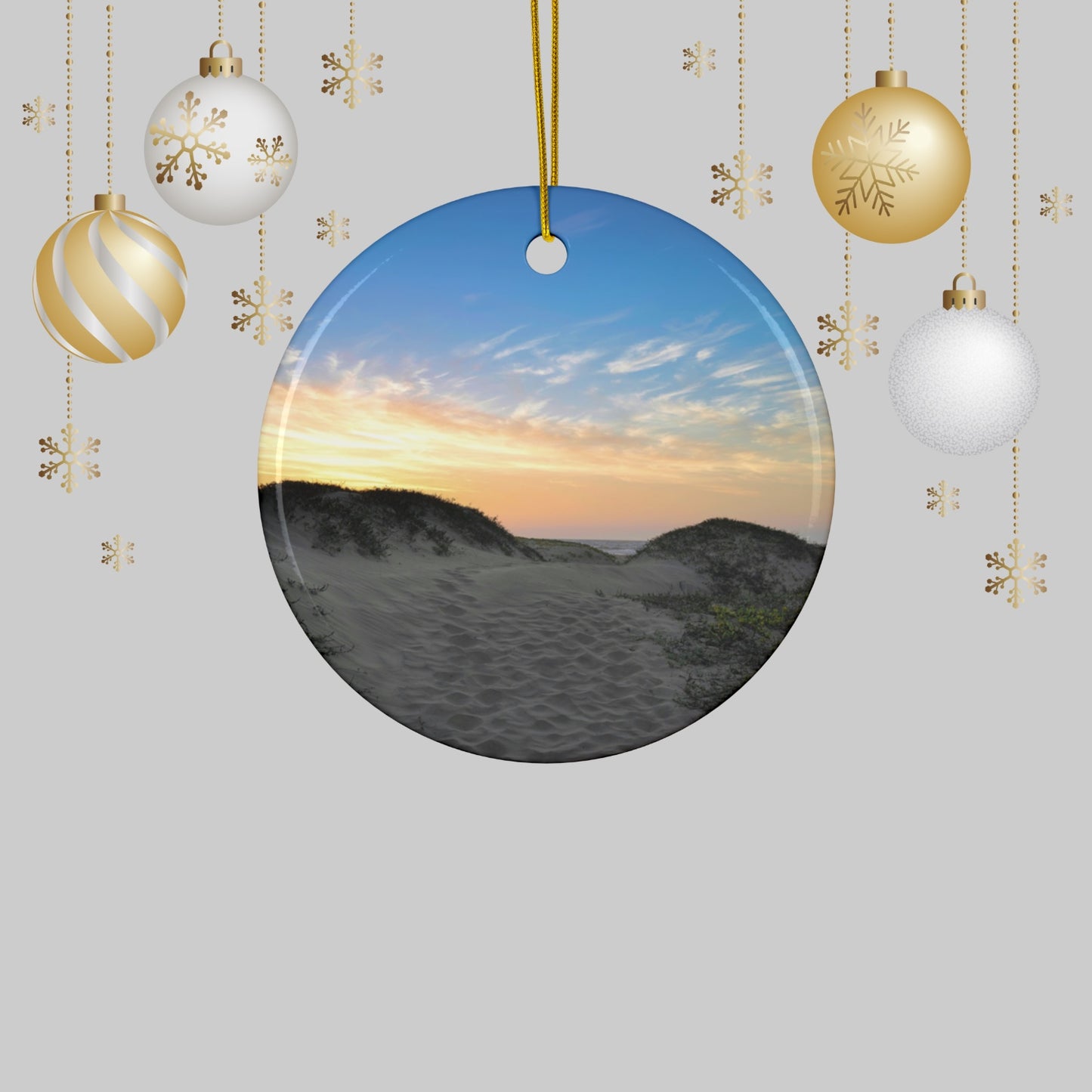 Ceramic holiday ornament featuring a landscape photograph of a California beach at sunset with rolling sand dunes, footprints, and gold-streaked clouds, surrounded by Christmas ornaments on a light gray background.