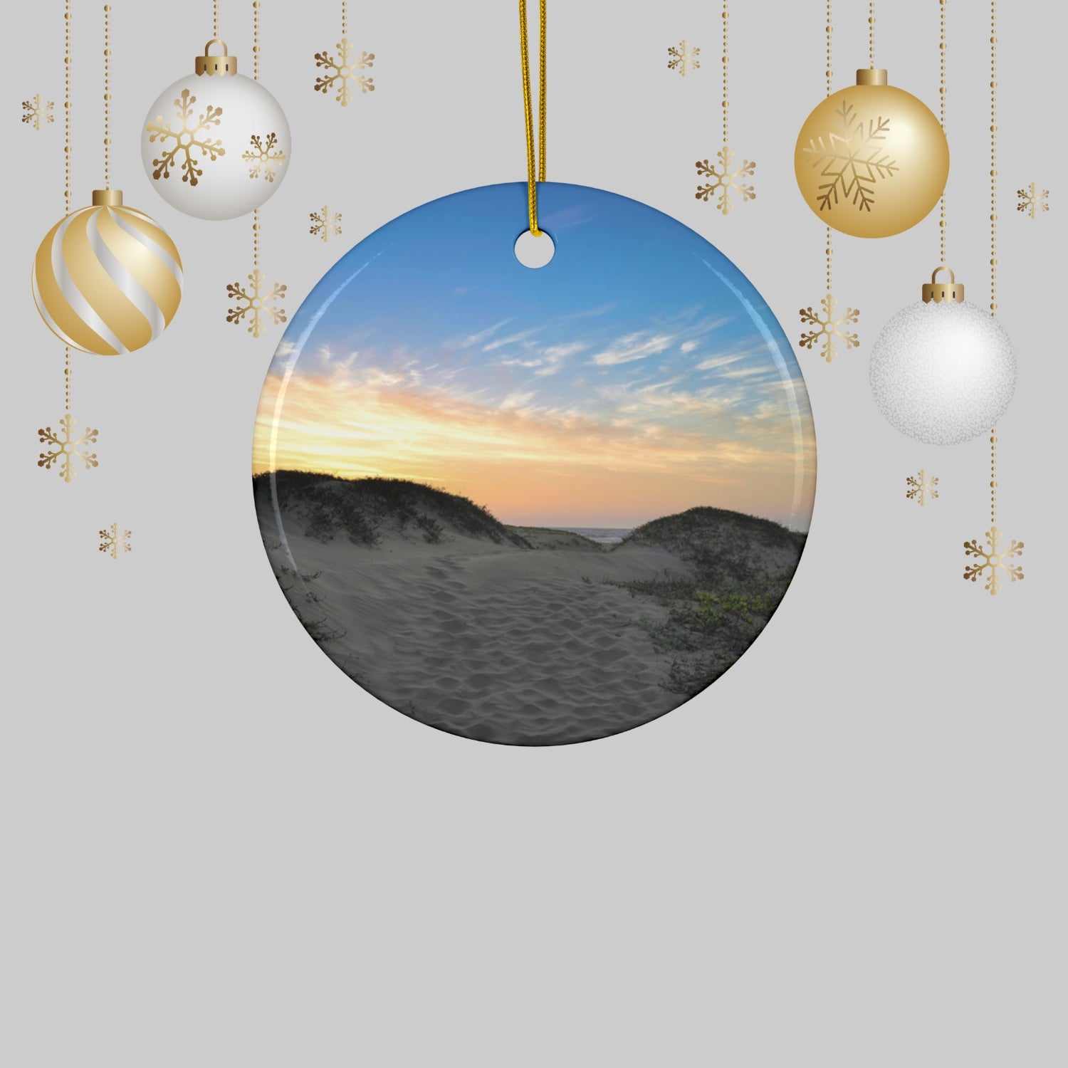 Ceramic holiday ornament featuring a landscape photograph of a California beach at sunset with rolling sand dunes, footprints, and gold-streaked clouds, surrounded by Christmas ornaments on a light gray background.