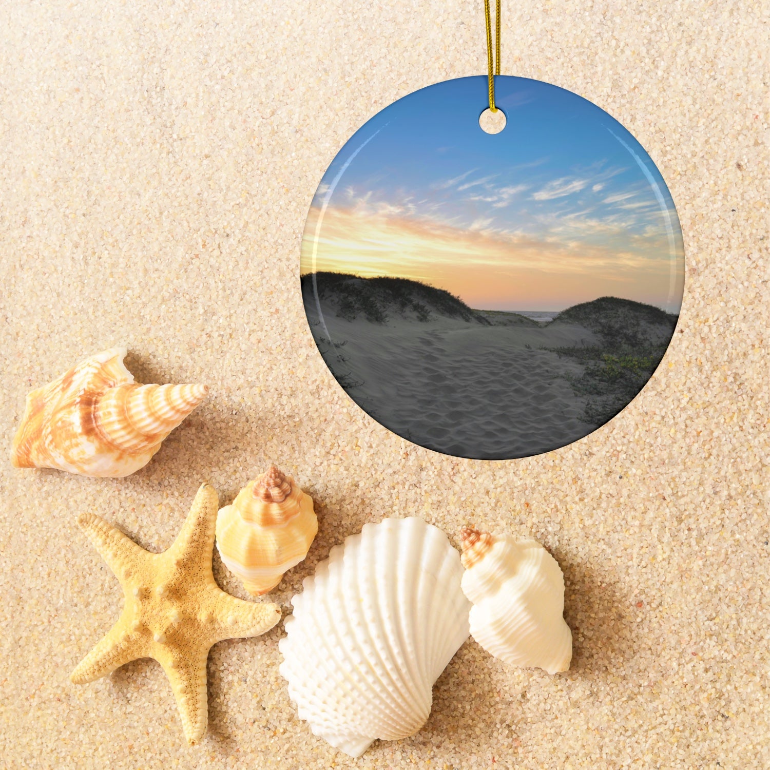 Ceramic holiday ornament featuring a landscape photograph of a California beach at sunset with rolling sand dunes, footprints, and gold-streaked clouds.on sand with shells