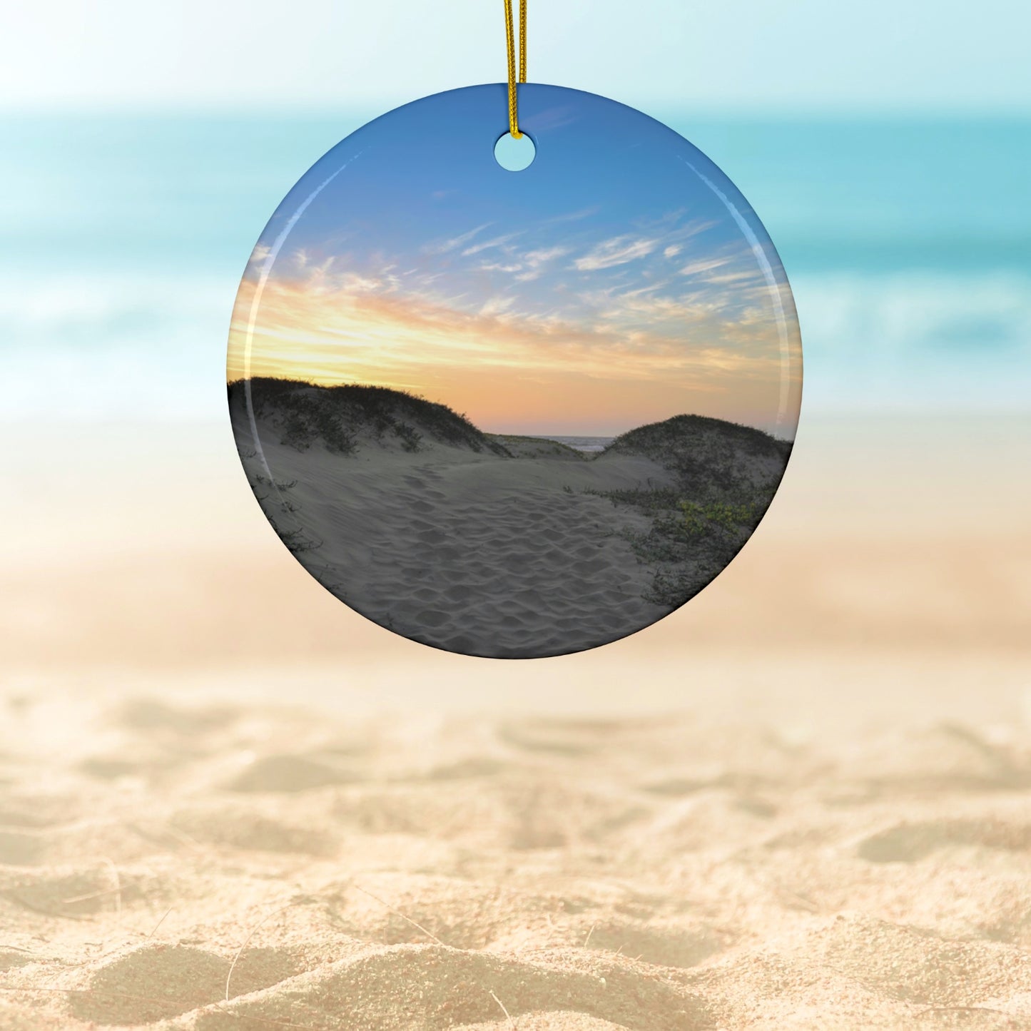 Ceramic holiday ornament featuring a landscape photograph of a California beach at sunset with rolling sand dunes, footprints, and gold-streaked clouds.