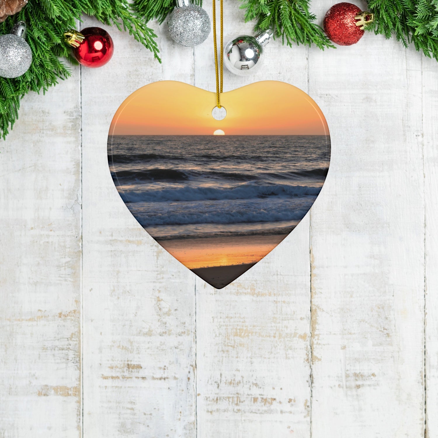 Heart-shaped ceramic Christmas ornament featuring a photograph of a vibrant amber and gold sunset over the Pacific Ocean with waves rolling toward the shore, hanging on a wooden surface with Christmas decorations.