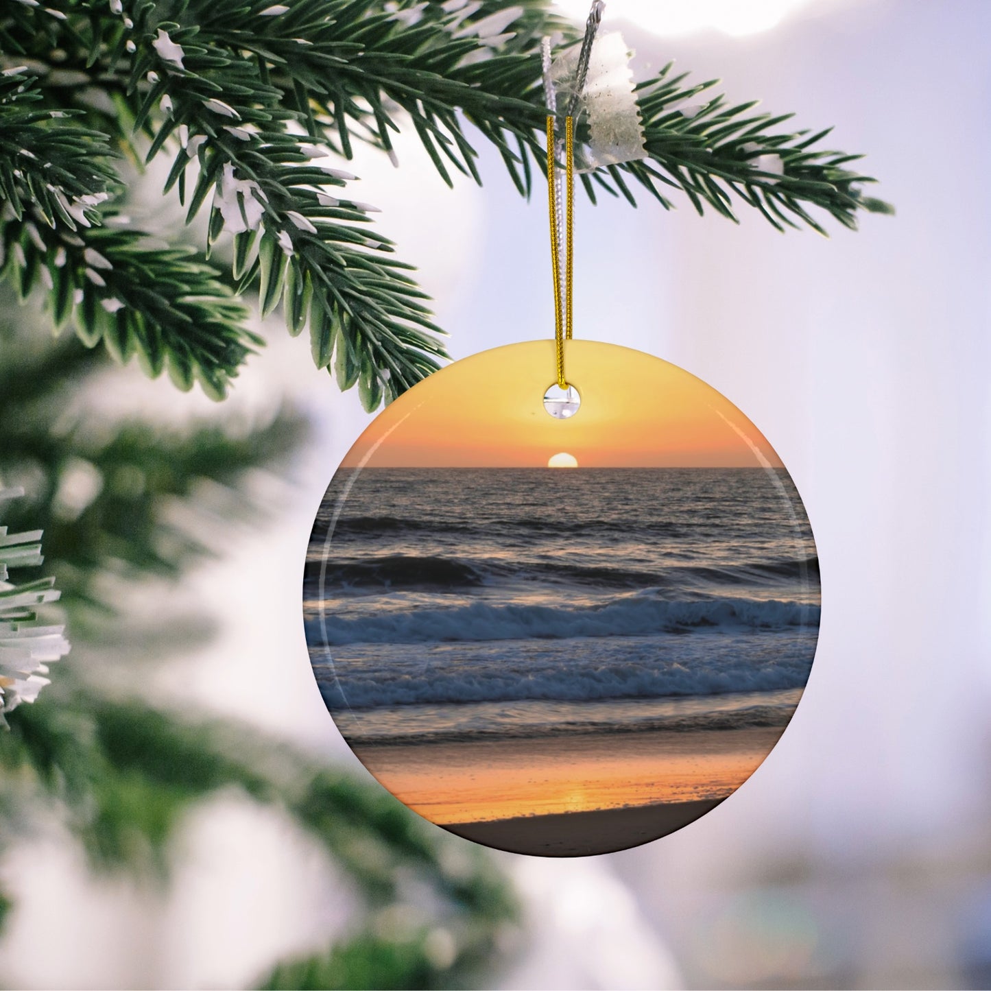 Ceramic Christmas ornament featuring a photograph of a vibrant amber and gold sunset over the Pacific Ocean with waves rolling toward the shore hanging on a Christmas tree branch.