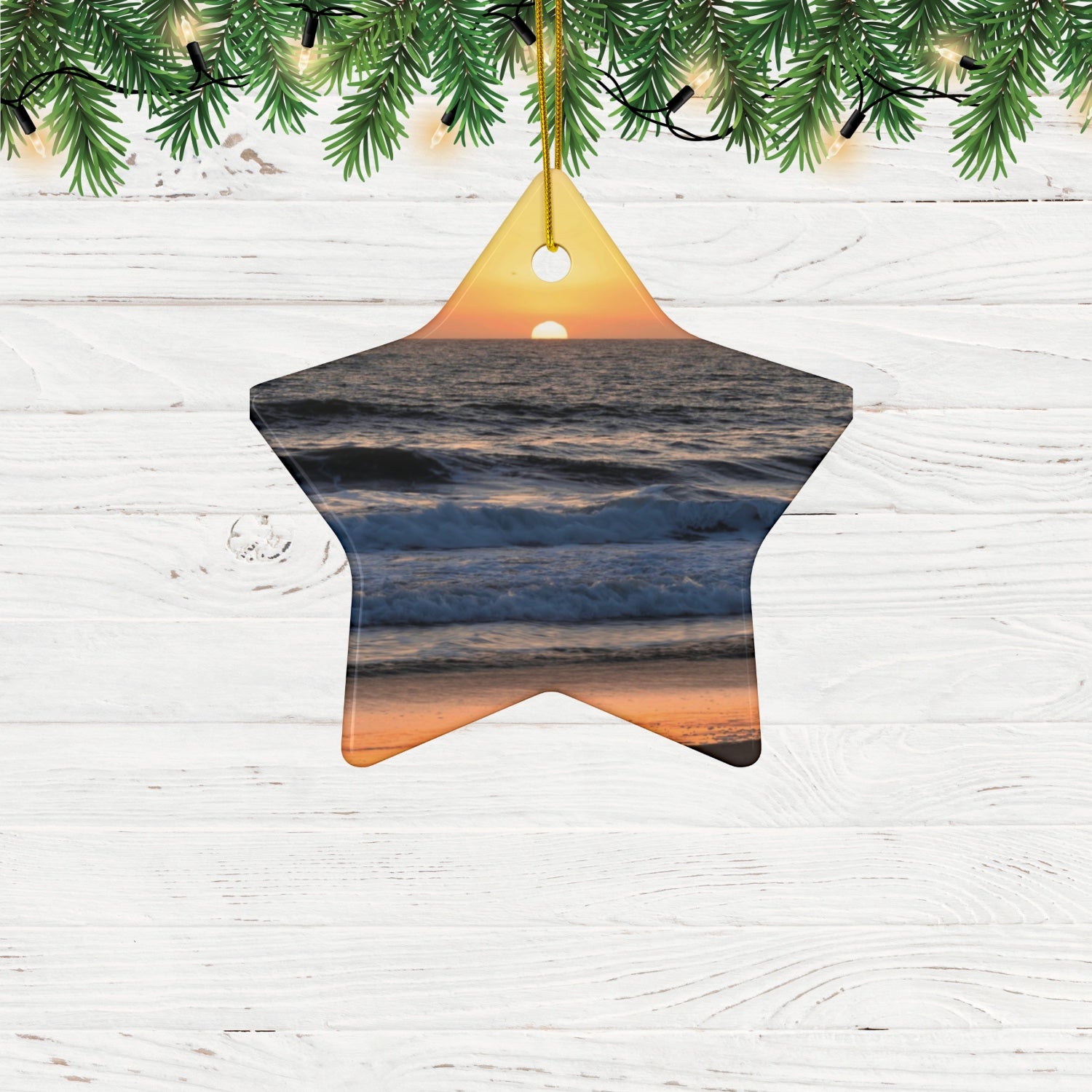 Star-shaped ceramic Christmas ornament featuring a photograph of a vibrant amber and gold sunset over the Pacific Ocean with waves rolling toward the shore on a white wooden background.