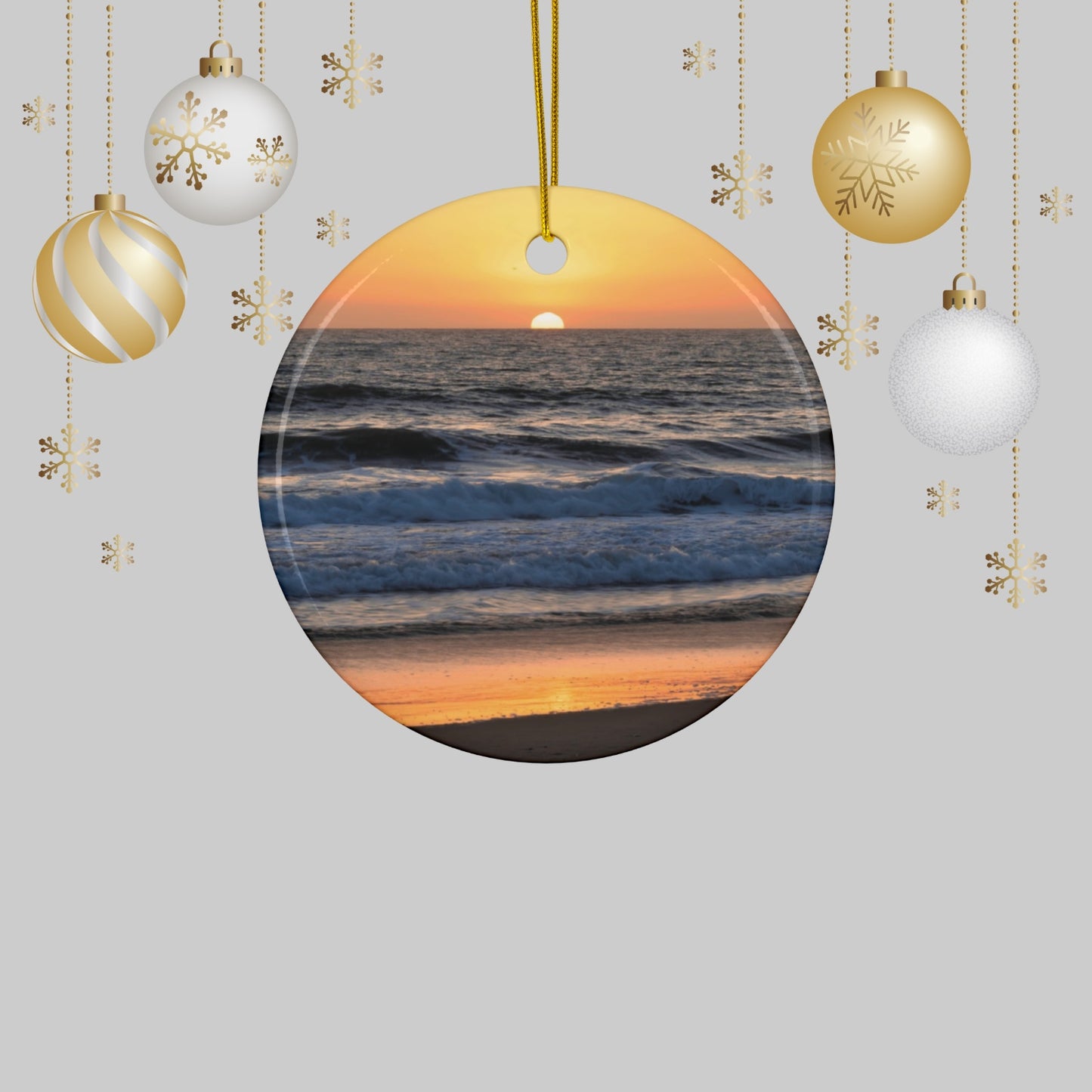 Ceramic Christmas ornament featuring a photograph of a vibrant amber and gold sunset over the Pacific Ocean with waves rolling toward the shore and Christmas ornaments on a light gray background.