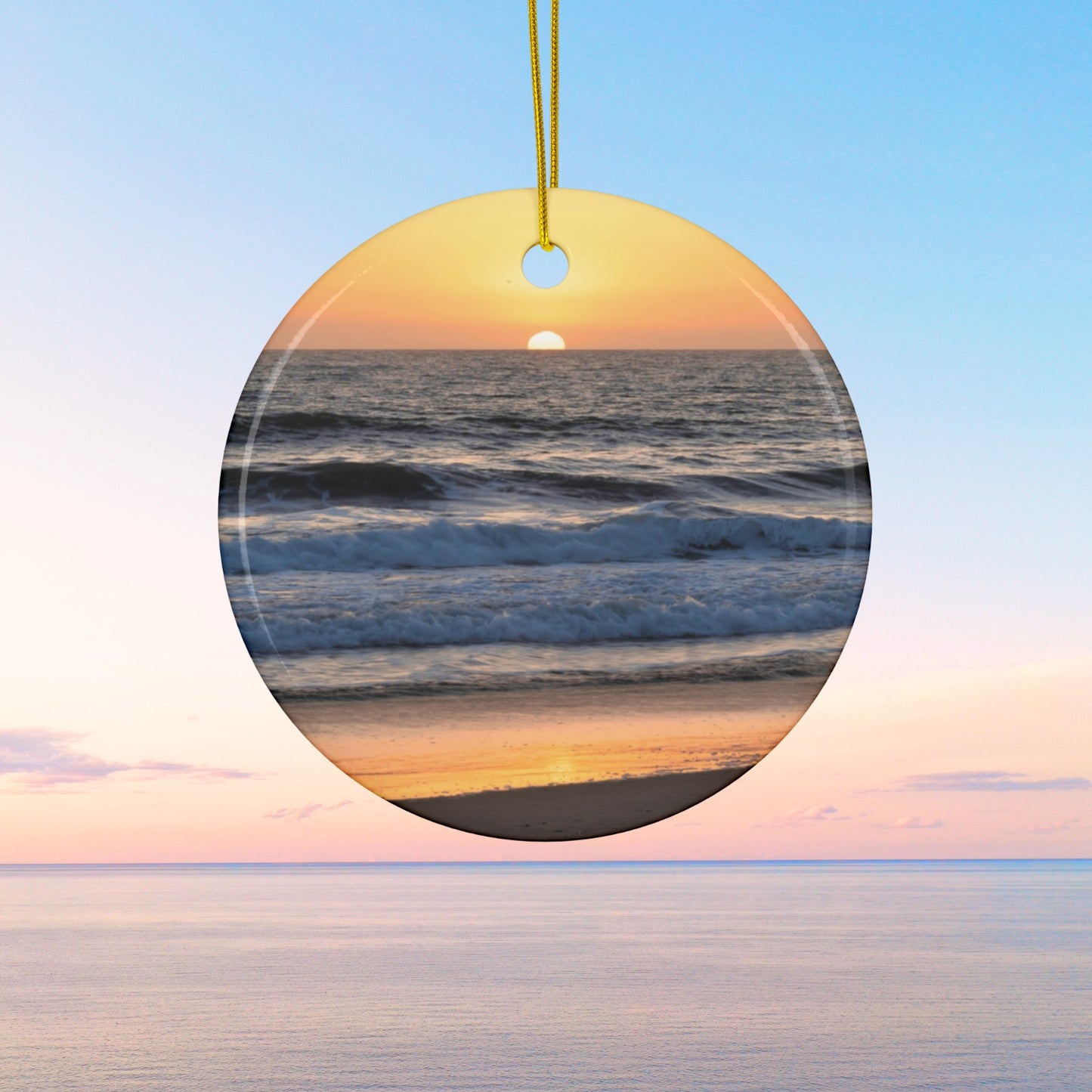 Ceramic Christmas ornament featuring a photograph of a vibrant amber and gold sunset over the Pacific Ocean with waves rolling toward the shore.